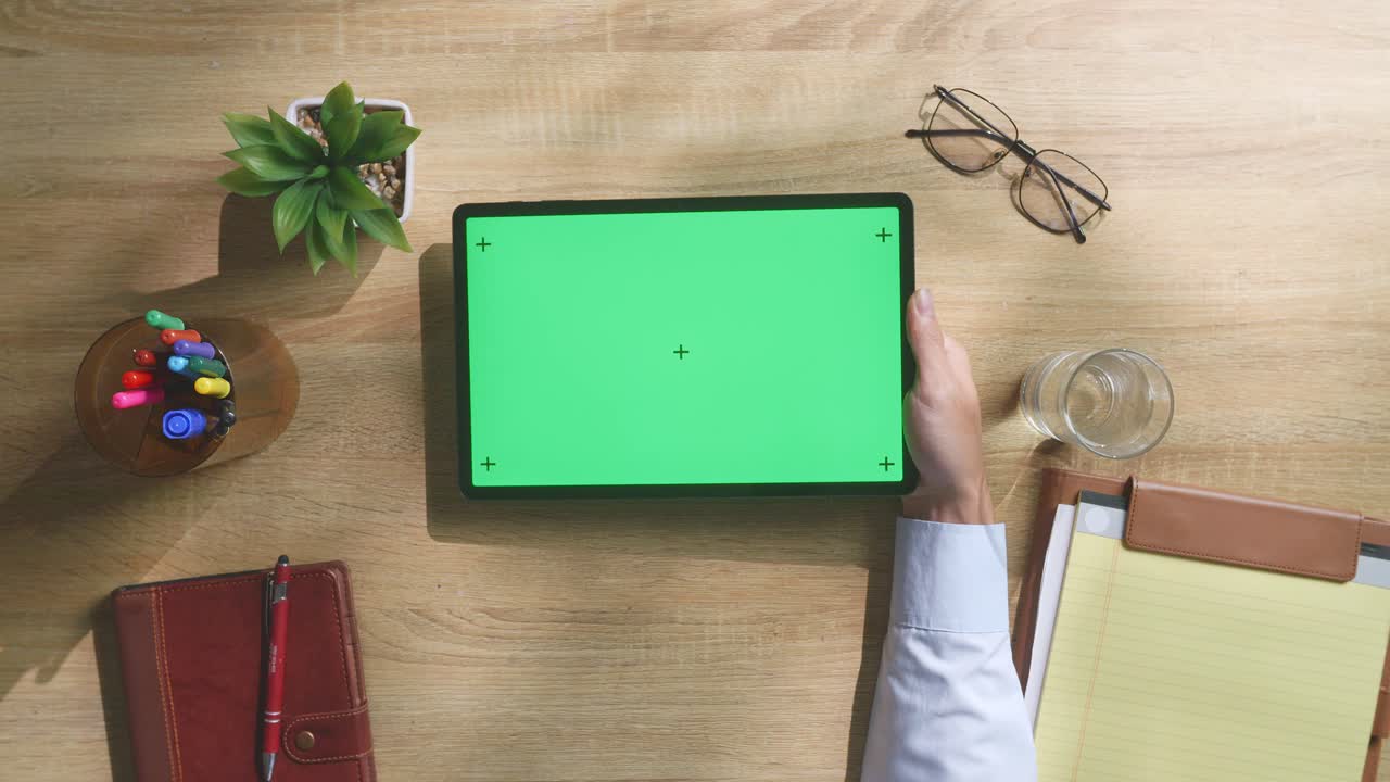 vista de arriba hacia abajo de una computadora portátil con pantalla verde simulada pantalla chromakey en un escritorio de oficina de madera al lado de la libreta con bolígrafos, gafas y un vaso de agua.