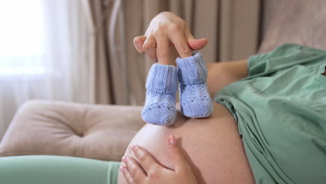 happy awaiting caucasian lady sitting on bed and enjoying maternity, copy space. young pregnant woman with baby booties at home