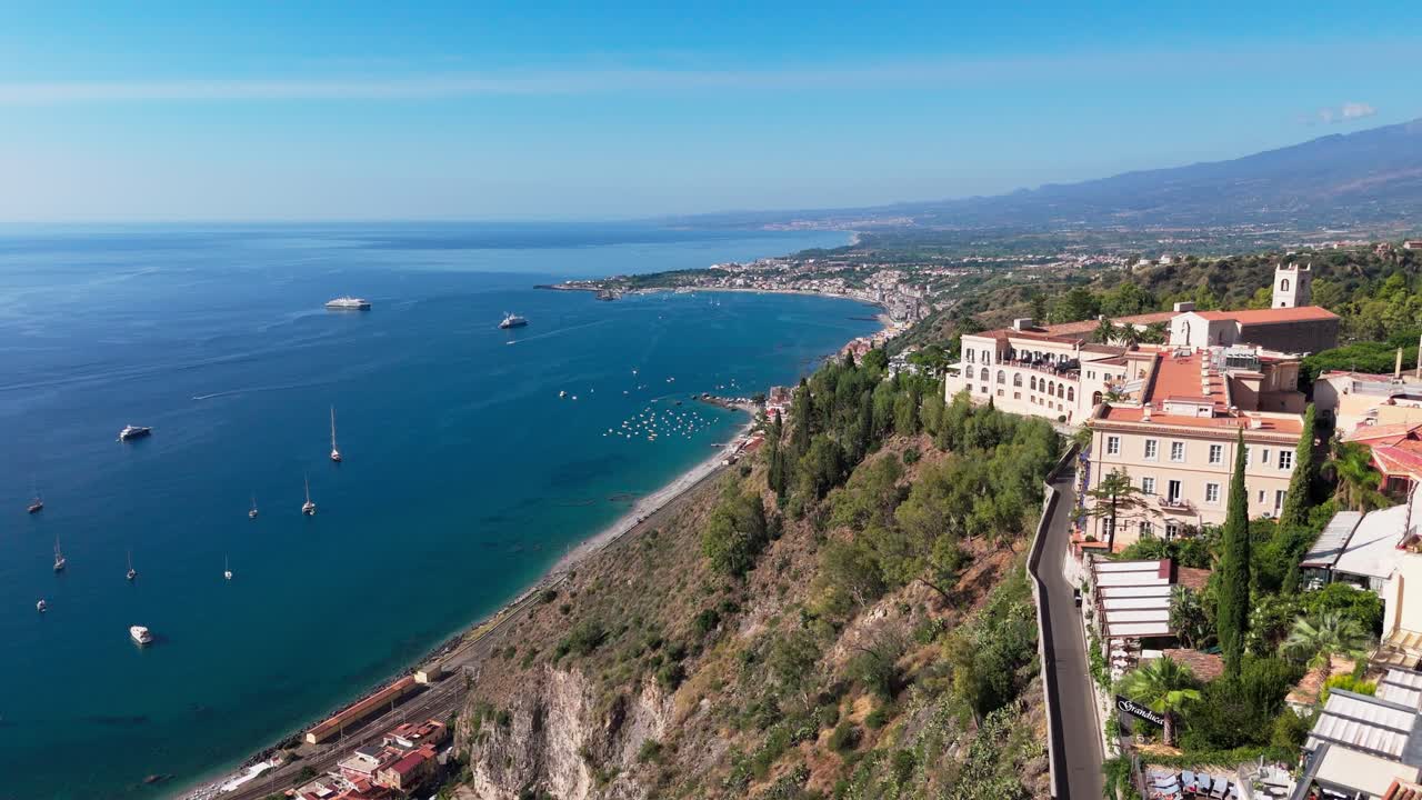 Aerial push-in shot featuring Taormina’s Duomo and Chiesa di San Giuseppe in the foreground, revealing the Naxos coastline and the sea beyond