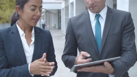 Front view of two business colleagues discussing project and watching presentation on tablet while walking at office building