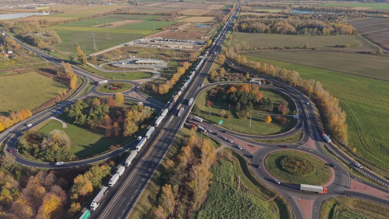 An aerial view of a busy highway interchange with multiple roundabouts and connecting roads. Trucks and cars move through the intersection surrounded by green fields and autumn trees