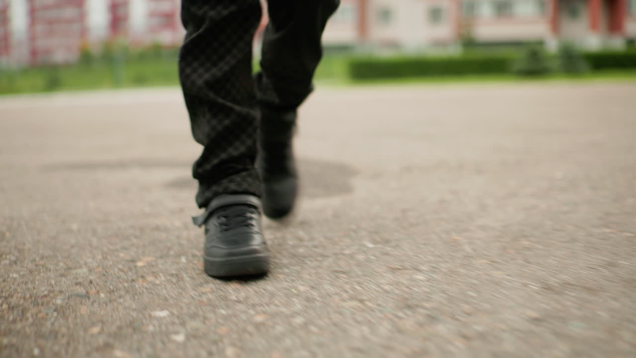 Vista de las piernas de un niño con zapatos negros caminando por una carretera asfaltada y acercándose a unas escaleras. El fondo desenfocado muestra edificios del campus y una barandilla de hierro, movimiento casual de estudiantes durante la rutina escolar diaria.