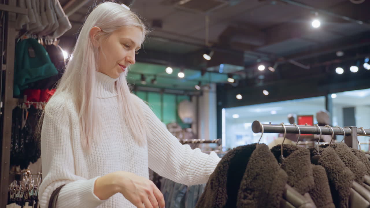 Smiling fashion lover with silver hair wearing white turtleneck sweater browses shearling jackets on boutique rack under spotlights, assessing fit, envisioning winter wardrobe update