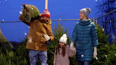 Glückliche-Familie-Kauft-Weihnachtsbaum-Auf-Dem-Markt.Familie,-Winterferien-Und-Menschenkonzept-–-Glückliche-Mutter,-Vater-Und-Kleine-Tochter-Kaufen-Weihnachtsbaum-Auf-Dem-Straßenmarkt-Am-Abend-Bei-Fallendem-Schnee