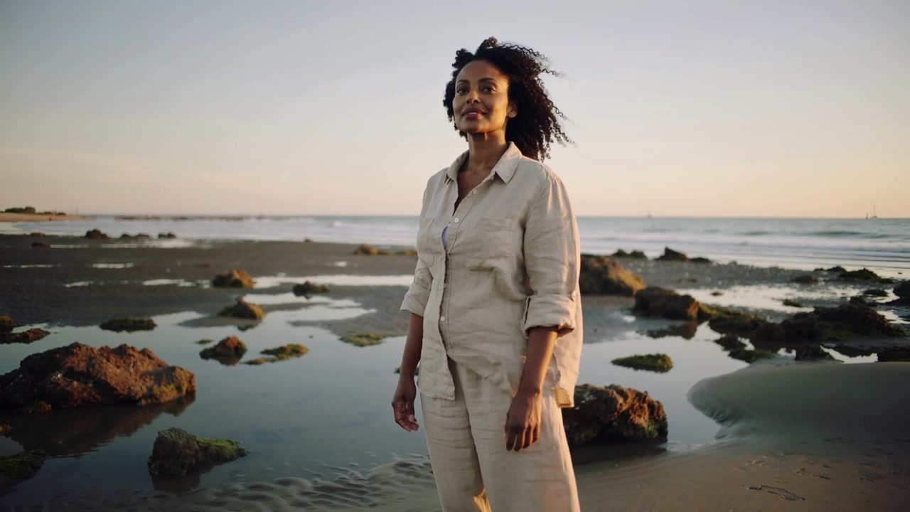Kvinde i beige linnedtøj vender sig og reagerer på kameraets tilgang på en stenet lavvandsstrand.