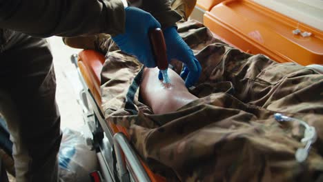 Close-up-of-a-confident-male-army-medic-in-a-dark-green-uniform-and-blue-gloves-inserting-a-needle-into-the-leg-of-an-unconscious-male-soldier-wounded-during-combat-in-the-ambulance-cabin