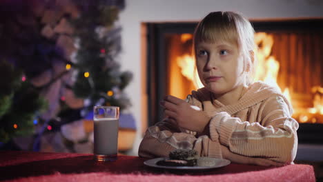 chica come galletas con leche en el fondo de la chimenea y el árbol de navidad