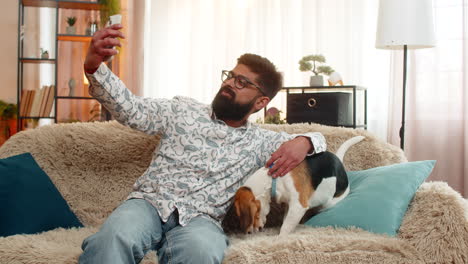 indian man joyfully takes selfie with beagle dog using smartphone while sitting on sofa at home