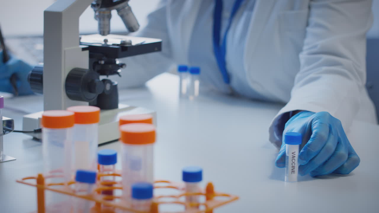 primer plano de un trabajador de laboratorio que lleva a cabo investigaciones utilizando un microscopio sosteniendo un tubo de ensayo con la vacuna etiquetada