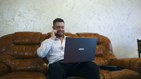 relaxed businessman is working in a hotel room on laptop. handsome man with laptop on knees speaks on the mobile phone.