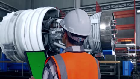 close up back view of asian male engineer with safety helmet working on a green screen tablet and looking around with airplane engine maintenance conducted, analytics checking the turbine