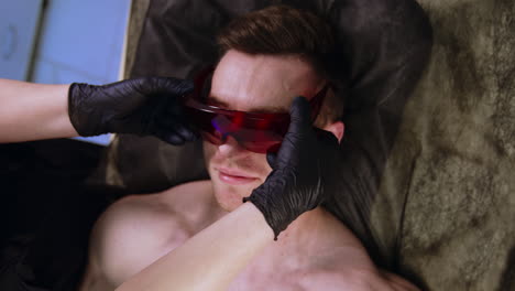 mid-aged caucasian man lies on the bed. specialist in latex gloves puts protective eyeglasses on a male client.