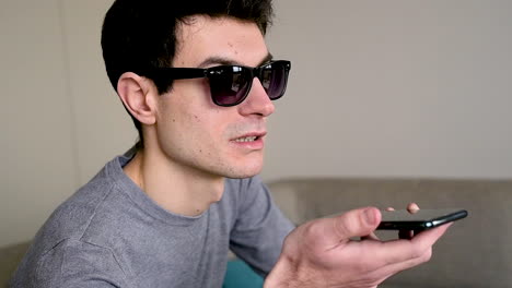 Close-Up-View-Of-A-Blind-Man-In-Sunglasses-Sitting-On-The-Sofa-At-Home-While-Holding-A-Smartphone-And-Having-A-Hands-Free-Call-1