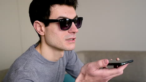 close up view of a blind man in sunglasses sitting on the sofa at home while holding a smartphone and having a hands free call 1