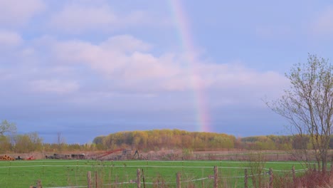 spring farm field at dusk with vivid rainbow and soft blue sky. trees sway gently in breeze, distant farm equipment adds rustic charm to the peaceful, pastoral landscape.