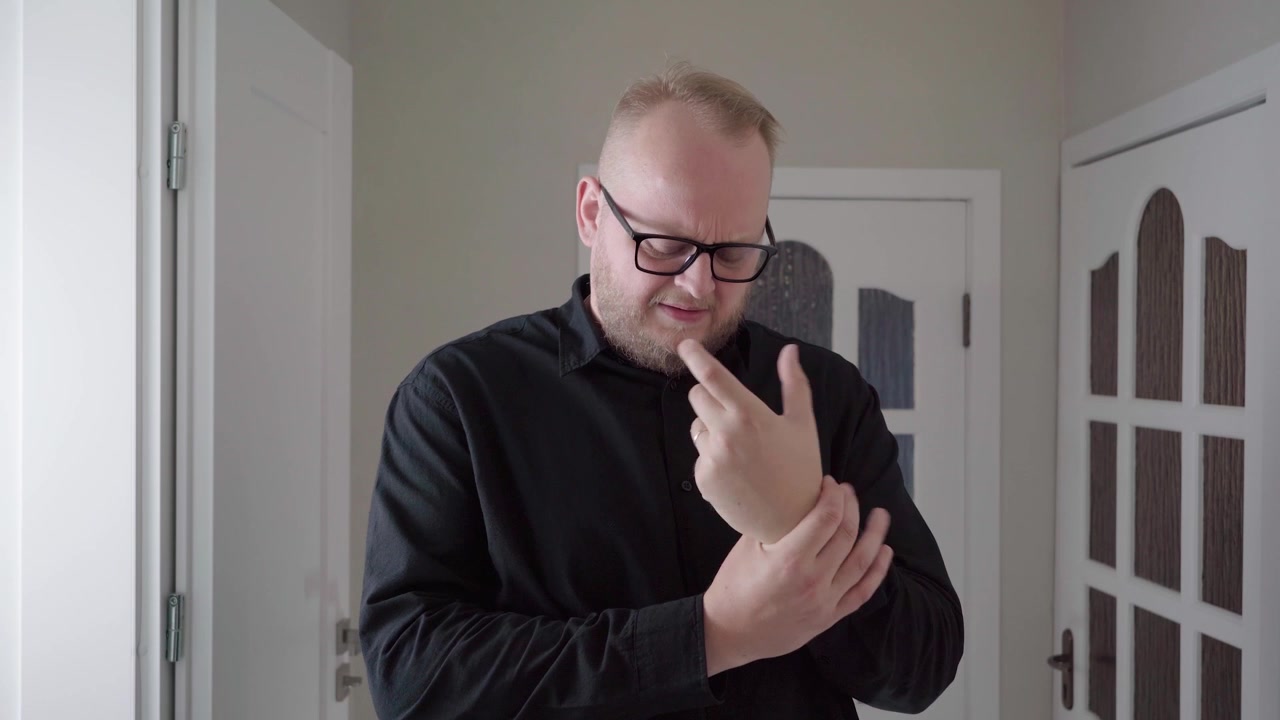 Adult man in a black shirt looks down and rubs his wrist and hand as if soothing joint pain in a bright home hallway.
