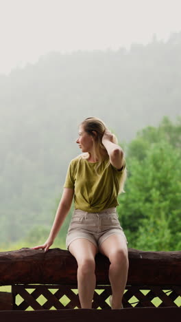 mujer atractiva con ropa cómoda toca el cabello sentada en una valla de troncos rústicos cerca del prado y la silueta de la montaña en un día de llovizna en cámara lenta