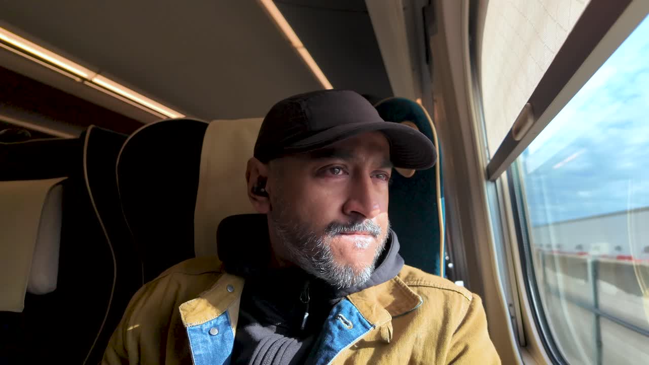Uomo sud-asiatico pensieroso che indossa auricolari e un berretto da baseball, guarda fuori dal finestrino mentre viaggia su un treno Shinkansen