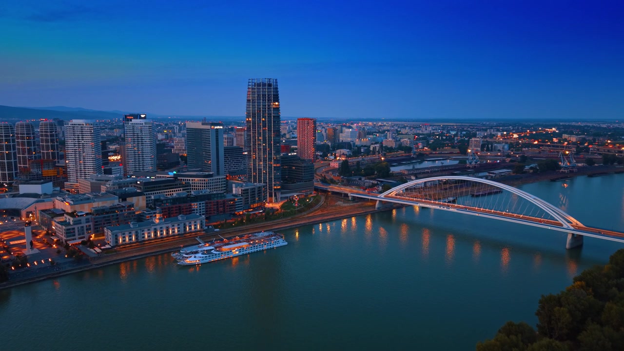 Moderno quartiere cittadino di Bratislava con il Ponte Apollo all'ora blu. Una ripresa aerea dall'alto rivela grattacieli illuminati, edifici sul lungofiume e una struttura ad arco durante la tarda serata