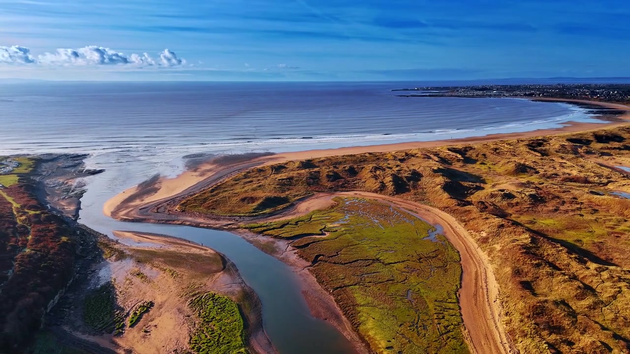 Vista Aérea de Foz de Rio Costeira com Dunas de Areia e Mar Aberto. Uma foz de rio encontra o mar aberto ao lado de extensas dunas de areia e um sapal verde com uma cidade portuária no horizonte distante.