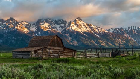 Hermoso-Timelapse-Dorado-Del-Amanecer-En-Una-Granja-Con-Granero-Y-Montaña-Nevada,-Nubes-Que-Fluyen-En-La-T