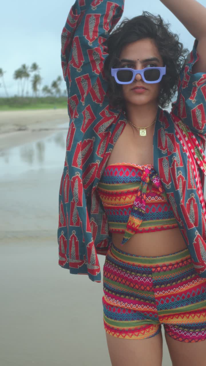 Stylish young woman with curly hair wearing colorful boho co ord set and blue sunglasses while posing on a beach, expressing confidence and a vibrant vacation lifestyle