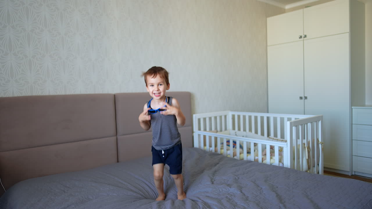 Kid wearing sleeveless t-shirt, shorts and sunglasses jumps on the double bed. Portrait of a happy child having fun at home.