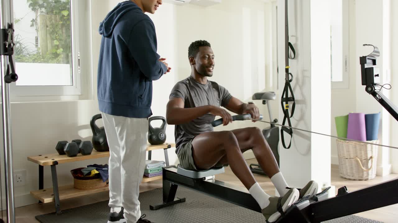 African American man rowing on machine after trainer pointing monitor for posture cues in home gym