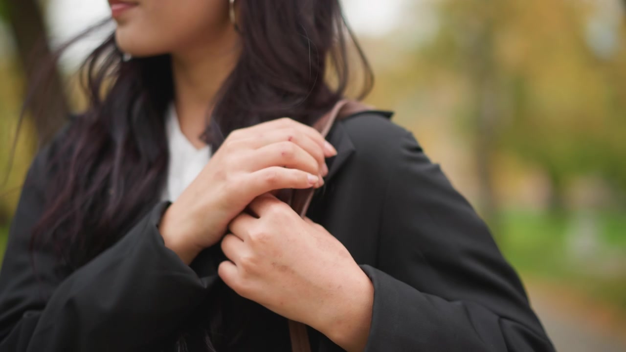 Primo piano di una donna che tiene e sistema una borsa tote in pelle marrone con la scritta FASHION in rilievo. Il soggetto indossa un cappotto nero, pantaloni bianchi e porta una borsa in un ambiente di parco all'aperto con luce naturale soffusa.