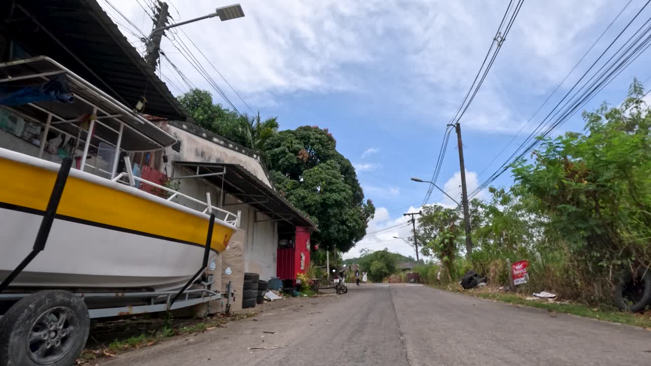 Low-angle camera glides along quiet rural road, passing boat, greenery, and roadside structures in daylight
