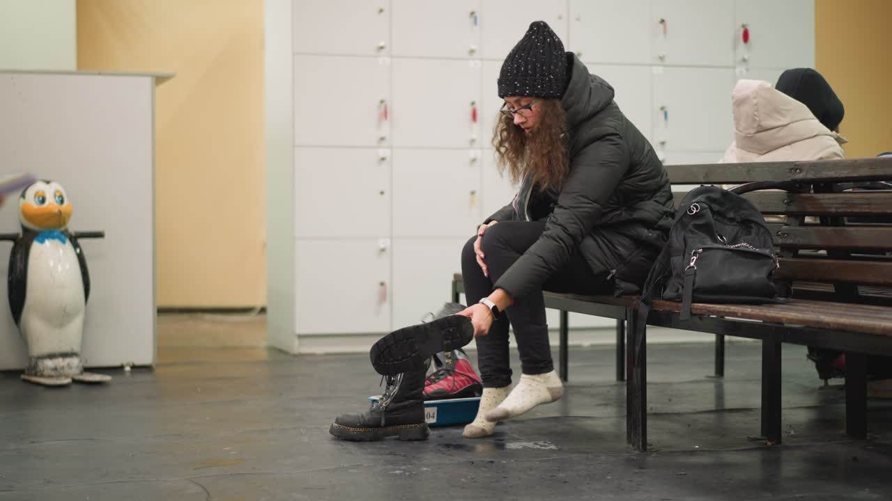 Female sitting on bench in locker room taking off black boots with chain detail while red and black ice skates rest inside blue tray on floor beside black backpack during winter preparation moment indoors