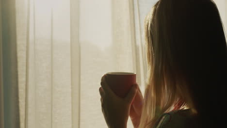 back view: a woman with a cup of hot tea looks out the window where the sun has risen. hot drink and good mood