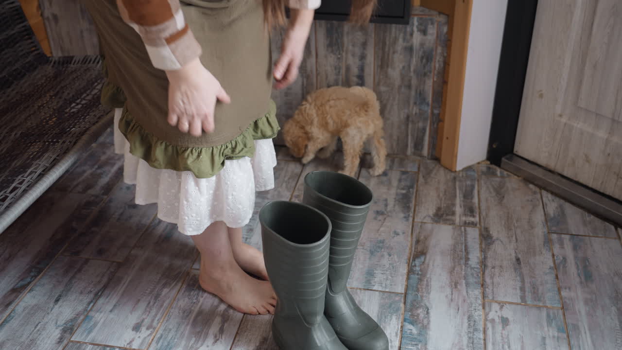 Owner standing barefoot on rustic wooden threshold holds black work boots near door as curious curly puppy with wagging tail moves around cozy indoor space under soft natural light