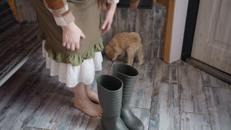 owner standing barefoot on rustic wooden threshold holds black work boots near door as curious curly puppy with wagging tail moves around cozy indoor space under soft natural light