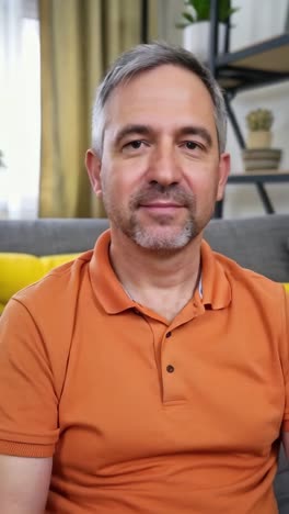 an adult man in an orange shirt is sitting on a couch. he has a beard and a mustache.