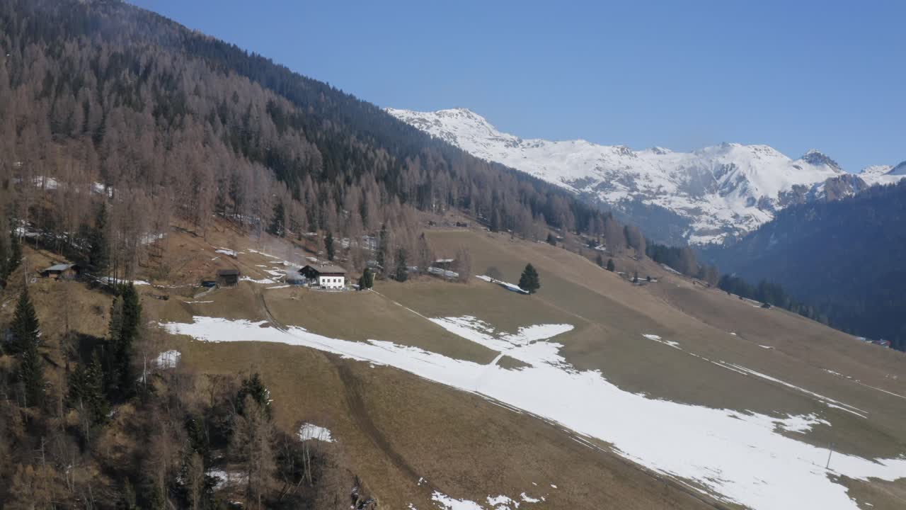 Drone flies over Navistal, Tyrol in spring, with snow on mountain peaks, a small hut in the valley, and patches of snow on the fields, capturing serene alpine beauty.