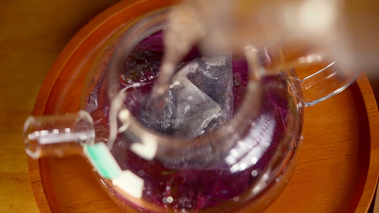 Top view of pouring hot water into transparent glass tea pot with tea bag and herbal leaves