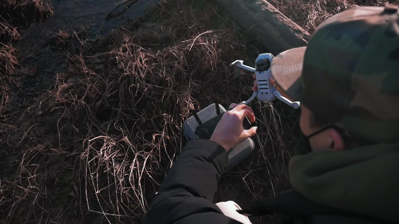 vista sobre el hombro de una persona que enciende su pequeño avión cuadricóptero dji mini drone uav con registros de barro de arena de hierba en el fondo