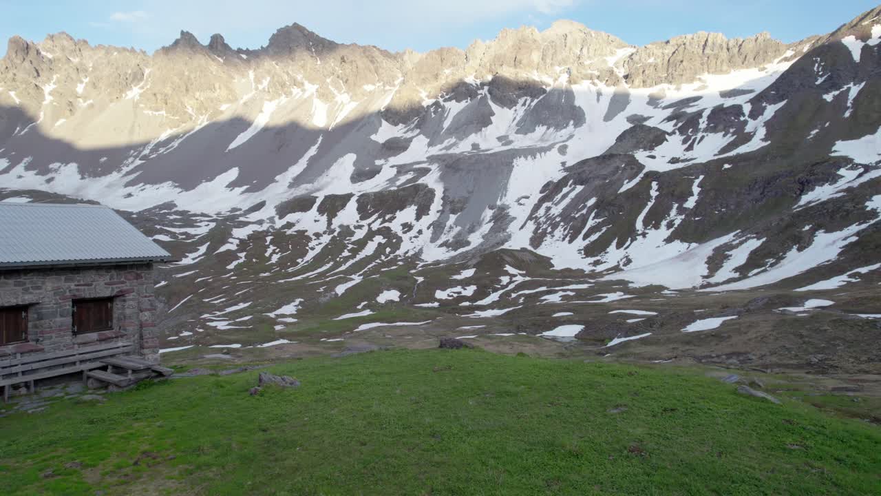 Aerial drone footage panning right from a Swiss alpine hut to reveal a dramatic, jagged mountain landscape with residual patches of snow and alpine meadows in Switzerland
