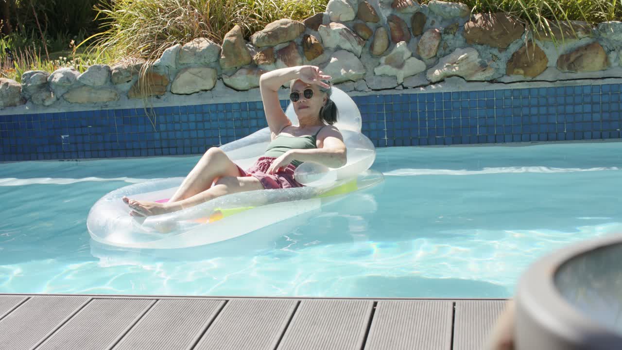 Senior woman is reclining on lounger in backyard pool, adjusting shades and shading eyes in sun