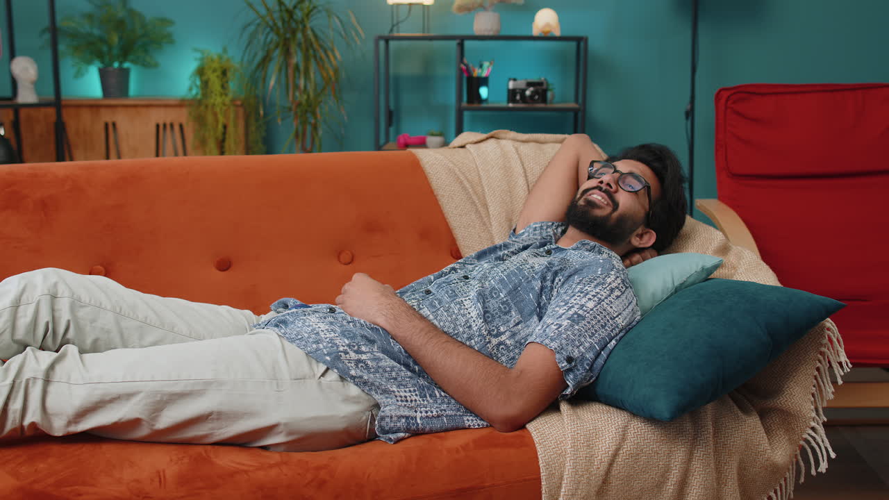 Tired indian man lying down in bed taking a rest at home napping falling asleep on sofa relaxing