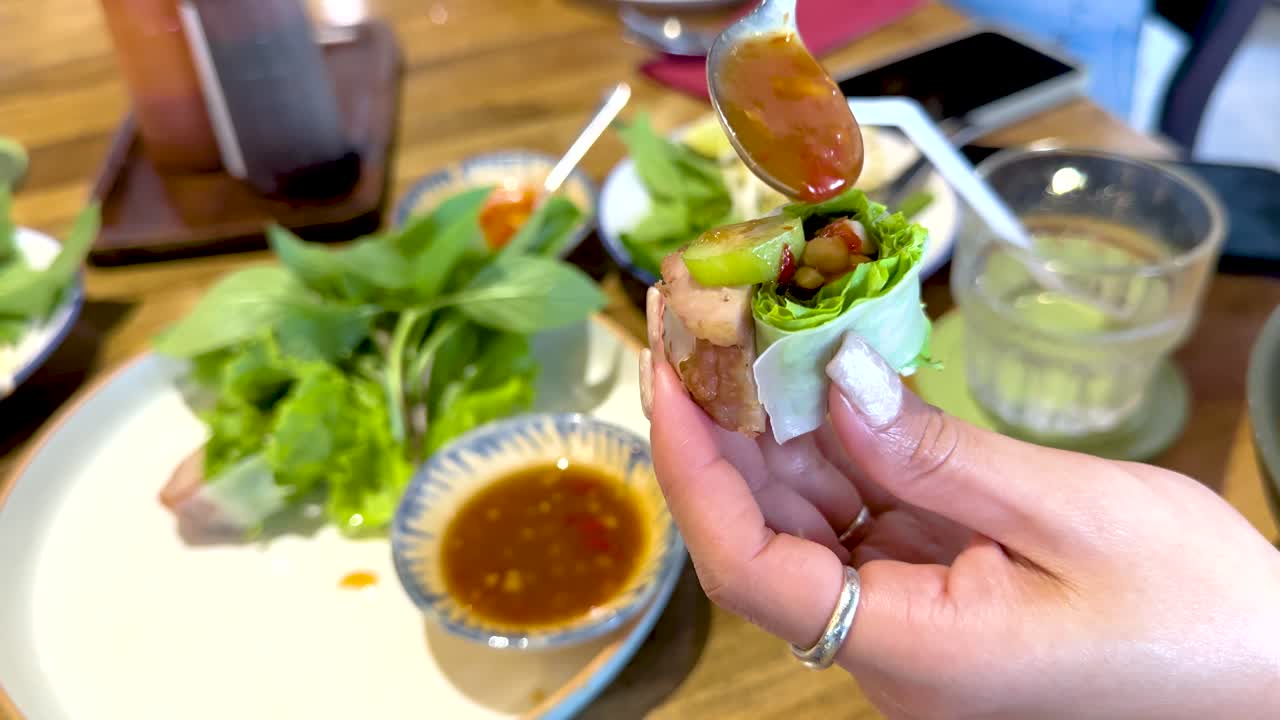 Hands assembling Vietnamese pork wraps with fresh greens and sauce in a well-lit Bangkok restaurant setting