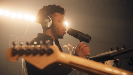 african american vocalist sings new composition into microphone in soundproof room. rock band on rehearsal in sound recording studio. man plays on guitar at background. concept of music production.