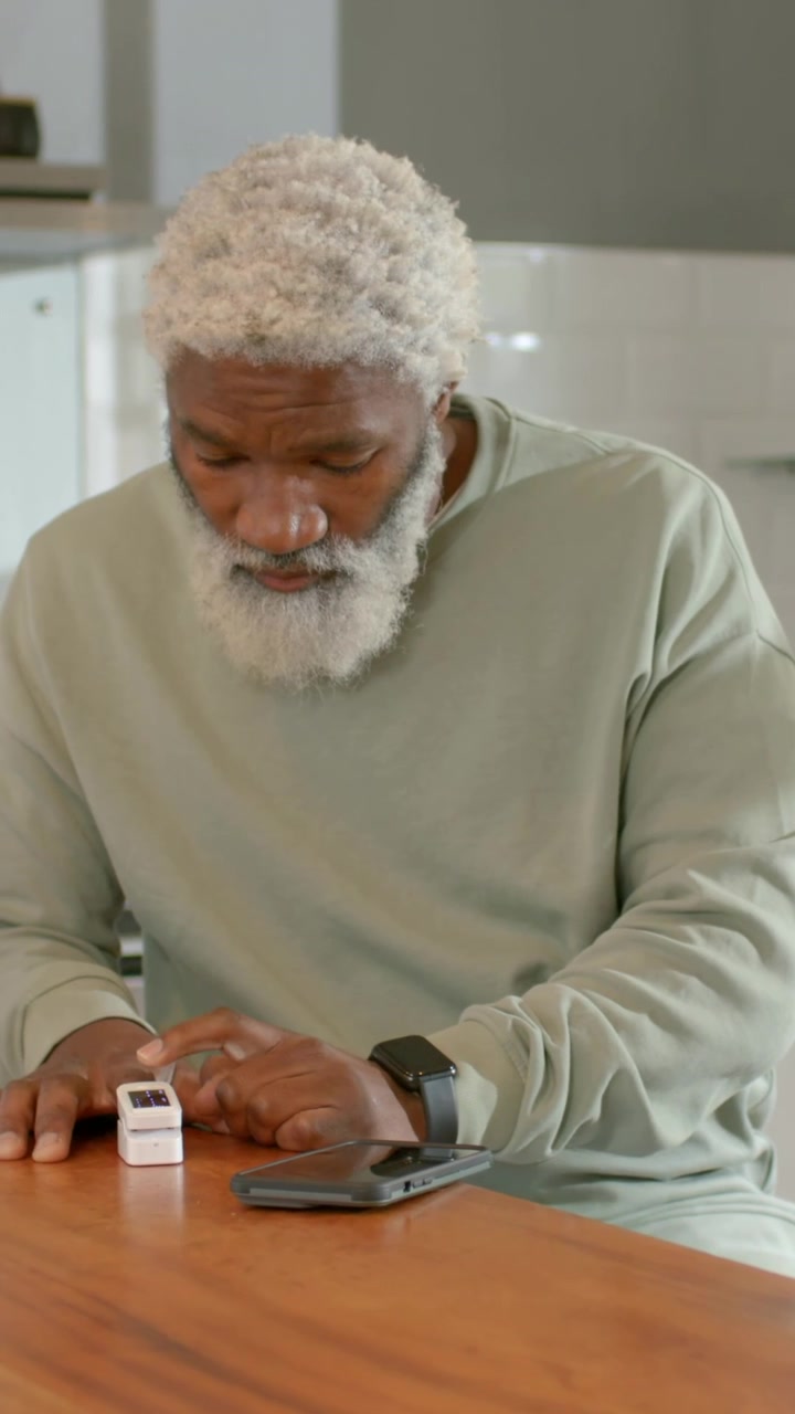 Vertical video: Leaning man in green top placing finger on fingertip device at home, checking phone