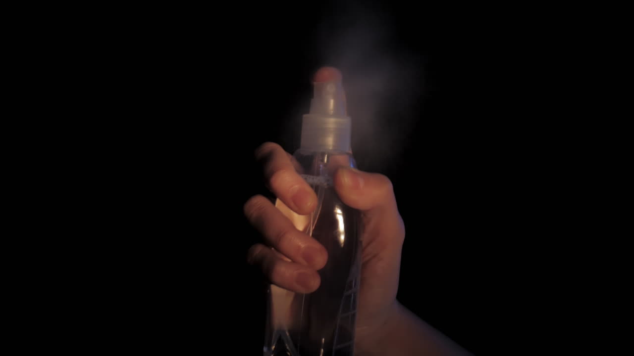 Mano rociando neblina de una botella de plástico transparente sobre fondo negro.
