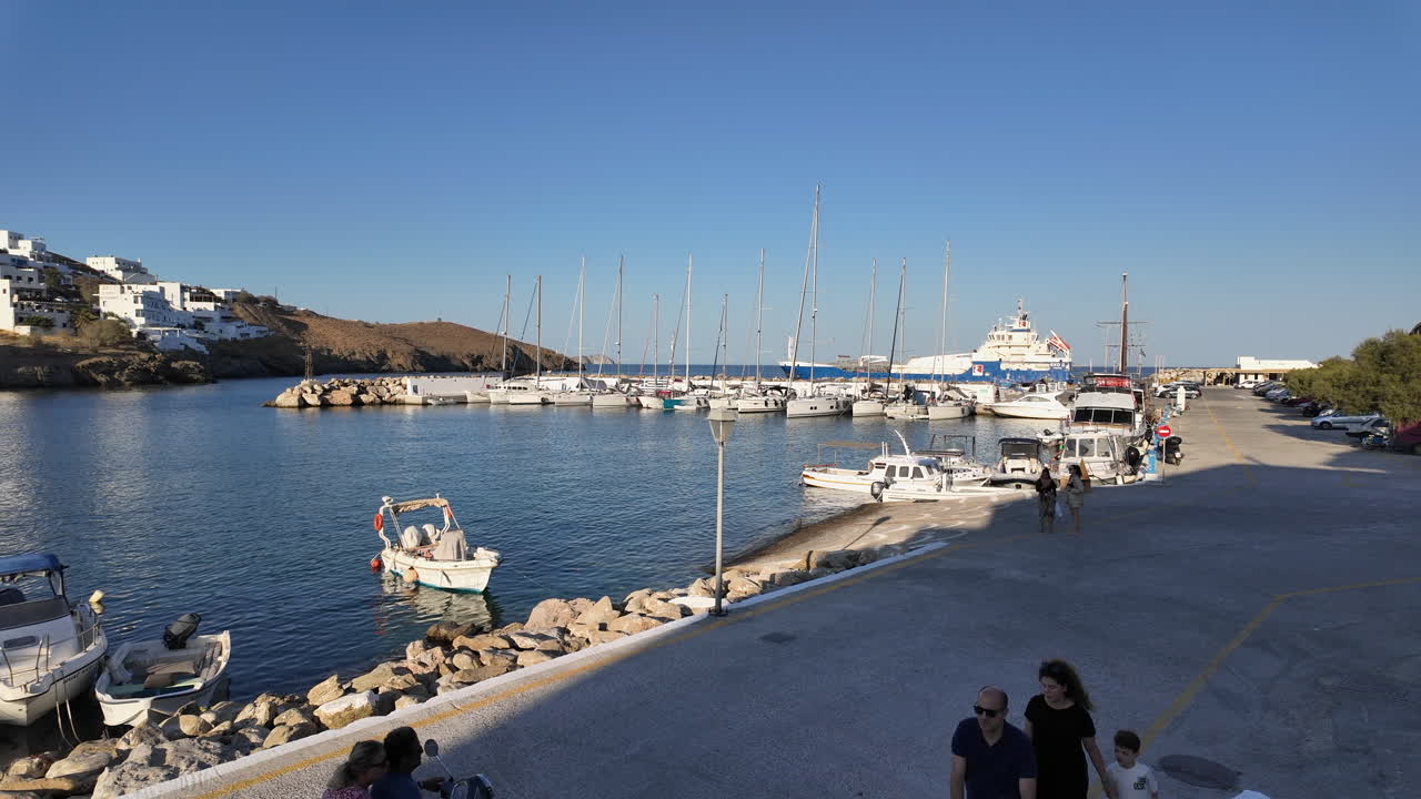 Town Quay of Astypalea on a sunny day where sail boats lined up and a big motor yacht across the harbor. people walk by and motorbikes passes.Beautiful white washed houses of the town at the scenery.