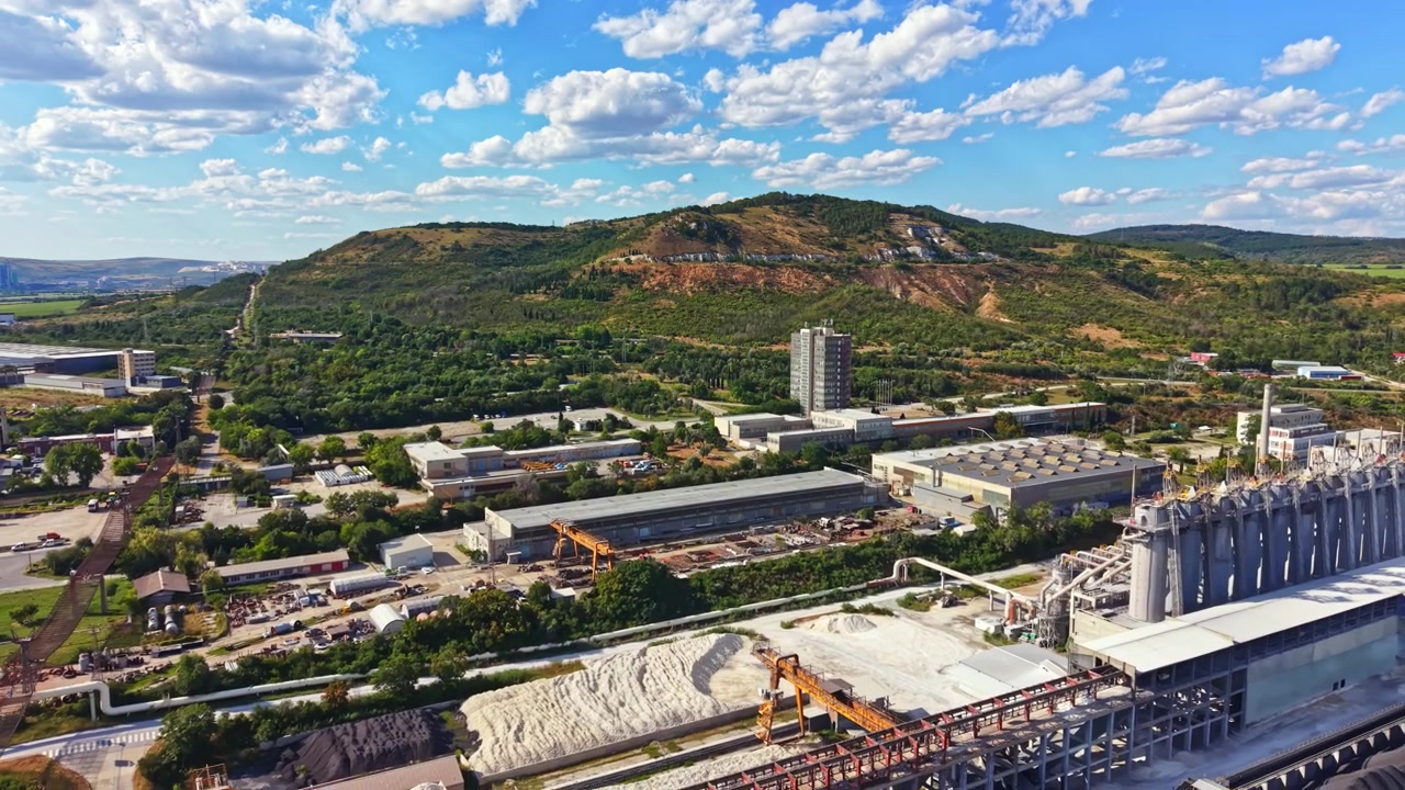 Utsikt over industriområde nær en åsside med bygninger og trær på en solrik dag med skyer på himmelen.