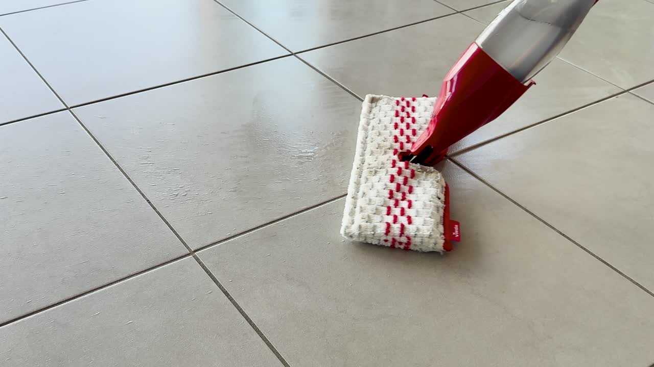 A person uses a red and white spray mop to clean large beige ceramic tiles in a sunlit, modern indoor space. Smooth camera movement follows the cleaning process