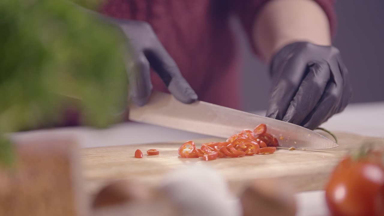 Slow-motion close-up of a chef slicing a red chili pepper on a wooden board. Black gloves ensure hygiene, while a sharp knife glides smoothly. Fresh ingredients like tomatoes and herbs add depth