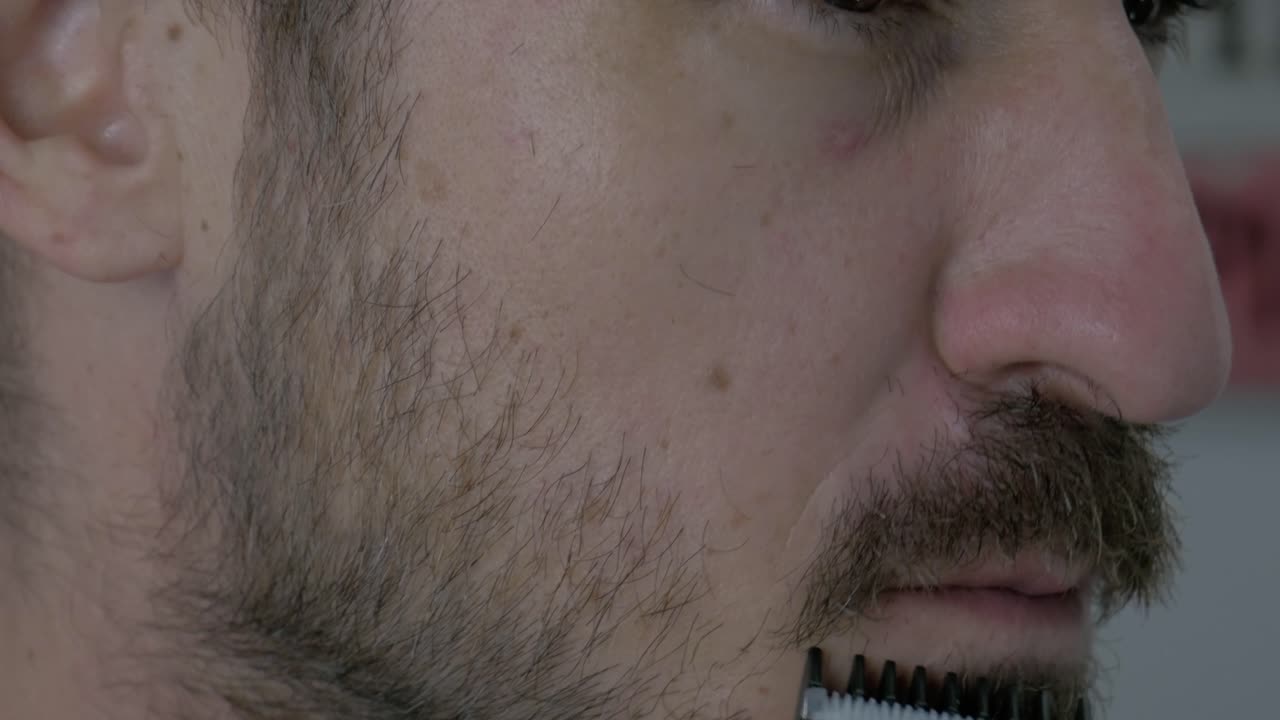 30대 백인 남자가 부드러운 머리카락 길이를 얻기 위해 전기 면도기로  ⁇ 수염을 자르고 있습니다.